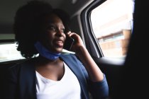 Mulher afro-americana usando máscara facial no carro falando em um smartphone. fora e sobre na cidade durante covid 19 coronavirus pandemia. — Fotografia de Stock