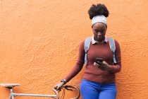 Portrait of african american woman using smartphone on street holding her bike out and about in the city during covid 19 coronavirus pandemic. — Stock Photo