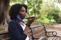 Femme afro-américaine portant un masque dans la rue assise sur un banc, parlant au téléphone. dehors et autour dans la ville pendant la pandémie de coronavirus covid 19. — Photo de stock