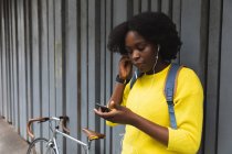 Mujer afroamericana usando teléfono inteligente en una calle, poniendo auriculares en sus oídos en la ciudad durante la pandemia covid 19 coronavirus. - foto de stock