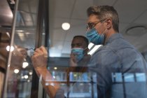 Caucasian man wearing face mask writing with marker pen on glass board at modern office. social distancing quarantine lockdown during coronavirus pandemic — Stock Photo