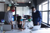 Diverse Kollegen tragen Gesichtsmasken mit Laptop, während sie während der Pandemie des Coronavirus 19 gemeinsam an moderner Bürohygiene und sozialer Distanzierung am Arbeitsplatz arbeiten. — Stockfoto
