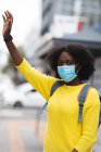 Porträt einer Afroamerikanerin mit Gesichtsmaske auf der Straße, die die Hand hebt. Während der 19-jährigen Coronavirus-Pandemie in der Stadt unterwegs. — Stockfoto