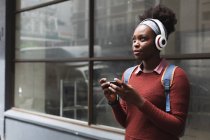 Retrato de una mujer afroamericana usando un smartphone en la calle, escuchando música con auriculares puestos. fuera de la ciudad durante la pandemia de coronavirus covid 19. - foto de stock