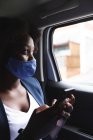 Mujer afroamericana con máscara facial en el coche usando un teléfono inteligente, mirando a través de una ventana. fuera de la ciudad durante la pandemia de coronavirus covid 19. - foto de stock