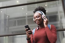 Porträt einer afrikanisch-amerikanischen Frau, die ihr Smartphone auf der Straße benutzt. Musik mit Kopfhörern hören. Während der 19-jährigen Coronavirus-Pandemie in der Stadt unterwegs. — Stockfoto