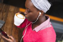 Африканская американка сидит в кафе, используя смартфон, пьет чашку кофе и слушает музыку. в городе во время 19 пандемических коронавирусов. — стоковое фото