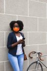 Mujer afroamericana que usa mascarilla facial usando smartphone en la calle en la ciudad durante la pandemia de coronavirus covid 19. - foto de stock