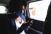 Mujer afroamericana con máscara facial en coche usando un teléfono inteligente en la ciudad durante la pandemia de coronavirus covid 19. - foto de stock
