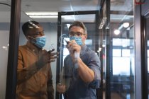 Белый мужчина в маске, пишет маркером на стеклянной доске в современном офисе. социальная изоляция от карантина во время пандемии коронавируса — стоковое фото