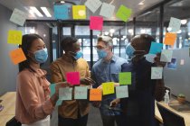 Разнообразные коллеги по офису в масках обсуждают заметки на стеклянной доске в современном офисе. социальная изоляция от карантина во время пандемии коронавируса — стоковое фото
