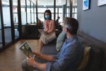 Mujer asiática que usa mascarilla facial usando tableta digital mientras el hombre caucásico sostiene el documento en la oficina moderna. distanciamiento social bloqueo de cuarentena durante la pandemia de coronavirus - foto de stock