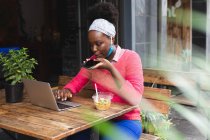 Femme afro-américaine assise dans un café à l'aide d'un ordinateur portable, parlant au téléphone et mangeant une salade dans la ville pendant la pandémie de coronavirus covid 19. — Photo de stock