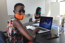 Портрет афро-американської жінки, одягненої в маску обличчя з використанням ноутбука в приміщенні для зустрічей в сучасному офісі. Відстань до карантину під час пандемії коронавірусу — стокове фото