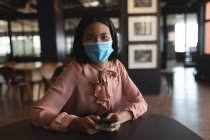 Портрет азійської жінки, одягненої в маску обличчя, тримає смартфон в сучасному офісі. Відстань до карантину під час пандемії коронавірусу — стокове фото