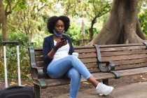 Femme afro-américaine portant un masque facial dans la rue assise sur un banc, en utilisant son smartphone. dehors et autour dans la ville pendant la pandémie de coronavirus covid 19. — Photo de stock