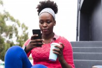 Donna afroamericana utilizzando smartphone e tenendo tazza in strada fuori e in giro per la città durante covid 19 pandemia coronavirus. — Foto stock