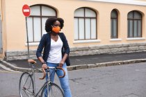 African american woman wearing face mask in street crossing the street and carrying her bicycle out and about in the city during covid 19 coronavirus pandemic. — Stock Photo