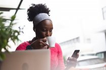 Африканская американка сидит в кафе, используя смартфон, пьет чашку кофе и слушает музыку. в городе во время 19 пандемических коронавирусов. — стоковое фото