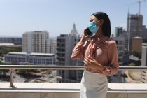 Mujer asiática con máscara facial hablando en el teléfono inteligente en la terraza en la oficina moderna. distanciamiento social bloqueo de cuarentena durante la pandemia de coronavirus - foto de stock