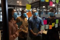 Разнообразные коллеги по офису в масках обсуждают заметки на стеклянной доске в современном офисе. социальная изоляция от карантина во время пандемии коронавируса — стоковое фото