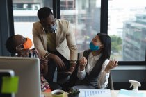 Diversos colegas con máscaras faciales riendo juntos en la oficina moderna. higiene y distanciamiento social en el lugar de trabajo durante la pandemia de coronavirus covid 19. - foto de stock