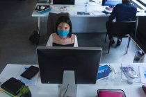 Вид зверху на азіатську жінку, яка носить маску для обличчя, використовуючи комп'ютер, сидячи на своєму столі в сучасному офісі. гігієна та соціальне дистанціювання на робочому місці під час пандемії коронавірусу 19 . — стокове фото