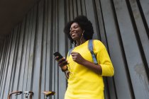 Mujer afroamericana usando smartphone en una calle escuchando música con auriculares. fuera de la ciudad durante la pandemia de coronavirus covid 19. - foto de stock