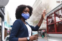 Mulher afro-americana usando máscara facial na rua segurando uma xícara de café e levantando a mão. fora e sobre na cidade durante covid 19 coronavirus pandemia. — Fotografia de Stock