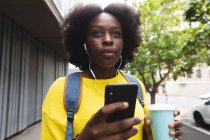 African american woman using smartphone on a street listening to music with earphones in. out and about in the city during covid 19 coronavirus pandemic. — Stock Photo
