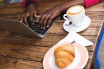 Femme afro-américaine assise dans un café à l'aide d'un ordinateur portable, buvant une tasse de café et mangeant un croissant dans la ville pendant la pandémie de coronavirus covid 19. — Photo de stock
