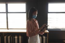 Mulher asiática usando máscara facial usando tablet digital no escritório moderno. bloqueio de quarentena por distanciamento social durante a pandemia do coronavírus — Fotografia de Stock