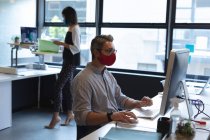 Белый мужчина в маске с помощью компьютера сидит на столе в современном офисе. социальная изоляция от карантина во время пандемии коронавируса — стоковое фото