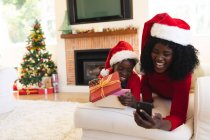 African american mother and daughter having video chat on tablet. wearing santa hats during christmas at home, girl is opening a gift. social distancing during covid 19 coronavirus quarantine lockdown — Stock Photo