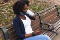 Afroamerikanerin mit Gesichtsmaske sitzt auf der Straße auf einer Bank und trinkt während der 19. Coronavirus-Pandemie in der Stadt eine Tasse Kaffee. — Stockfoto