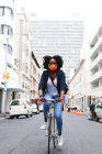 African american woman wearing face mask in street riding a bicycle out and about in the city during covid 19 coronavirus pandemic. — Stock Photo