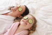 Donna e figlia caucasica si divertono in camera da letto. mettendo fette di cetriolo sugli occhi. godendo di tempo di qualità a casa durante coronavirus covid 19 isolamento pandemico. — Foto stock
