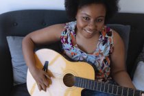 Mujer afroamericana sonriente sentada en un sofá tocando la guitarra acústica. autoaislamiento hobby tiempo música en casa durante coronavirus covid 19 pandemia. - foto de stock