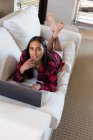 Mixed race woman lying on couch at home using laptop and smiling. self isolation during covid 19 coronavirus pandemic. — Stock Photo