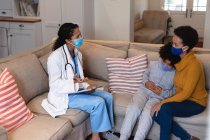 Mestiça mãe e filha conversando com mestiça médica sentada no sofá. auto-isolamento em casa juntos durante coronavírus covid 19 pandemia. — Fotografia de Stock