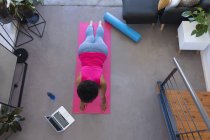 Mujer afroamericana usando laptop haciendo ejercicio usando ropa deportiva. auto aislamiento fitness tecnología comunicación en el hogar durante coronavirus covid 19 pandemia. - foto de stock