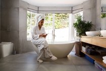 Femme de race mixte portant un peignoir assis sur la baignoire à l'aide d'un smartphone. auto-isolement à la maison pendant la pandémie de coronavirus covid 19. — Photo de stock