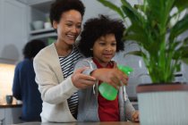 Mixed race woman and daughter watering plants in kitchen. self isolation quality family time at home together during coronavirus covid 19 pandemic. — Stock Photo