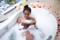 Смешанная расовая женщина лежит в ванне расслабляясь и читая книгу. самоизоляция во время пандемии 19 коронавируса. — стоковое фото