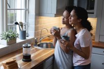 Sonriente pareja de lesbianas de raza mixta bebiendo café y abrazándose en la cocina. autoaislamiento calidad tiempo en casa juntos durante coronavirus covid 19 pandemia. - foto de stock