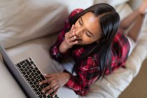 Mixed race woman lying on a couch at home using laptop and smiling. self isolation during covid 19 coronavirus pandemic. — Stock Photo