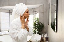 Donna razza mista guardando allo specchio applicando crema viso in bagno. auto isolamento a casa durante covid 19 coronavirus pandemia. — Foto stock