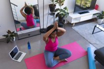 African american woman doing yoga meditation sitting on mat wearing sports clothes. laptop in the background. self isolation fitness wellbeing technology at home during coronavirus covid 19 pandemic. — Stock Photo