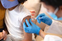 Chica de raza mixta con máscara facial que recibe la vacuna contra la gripe por parte de una doctora. autoaislamiento en casa durante la pandemia del coronavirus covid 19 - foto de stock