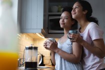 Усміхнена змішана гонка лесбійська пара п'є каву і приймає на кухні. Якість самоізоляції вдома разом під час пандемії коронавірусу 19 . — стокове фото
